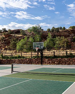 An outdoor sports court with a basketball hoop and tennis court, set against a backdrop of hills and a partly cloudy sky.