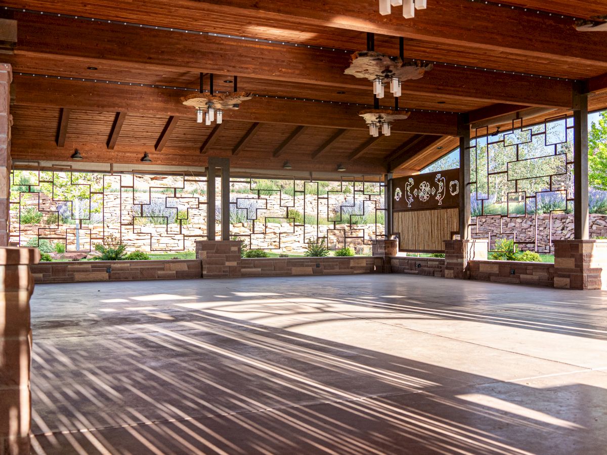 The image shows an open, covered pavilion with wooden beams and stained glass windows, allowing natural sunlight to filter through.