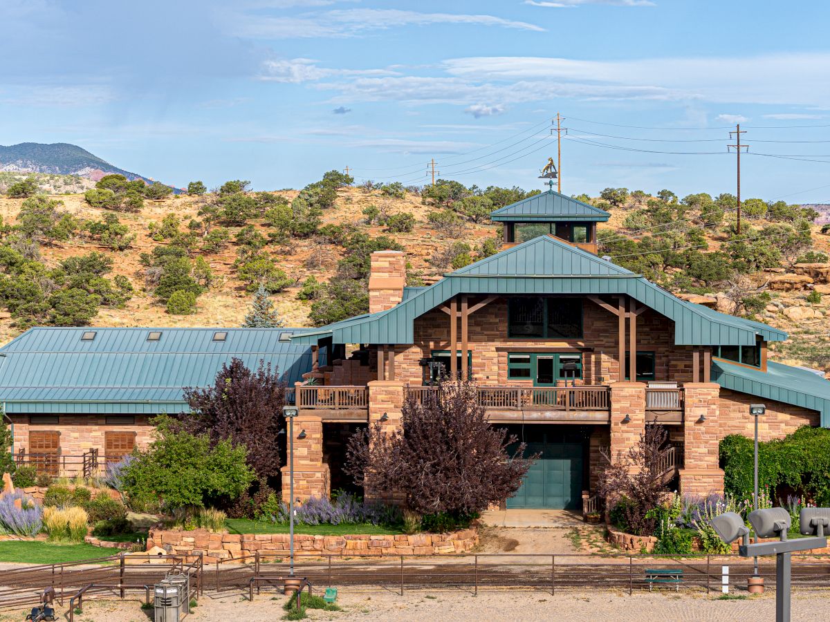 A large, rustic house with a green roof sits in a desert landscape, surrounded by trees and shrubs under a blue sky.