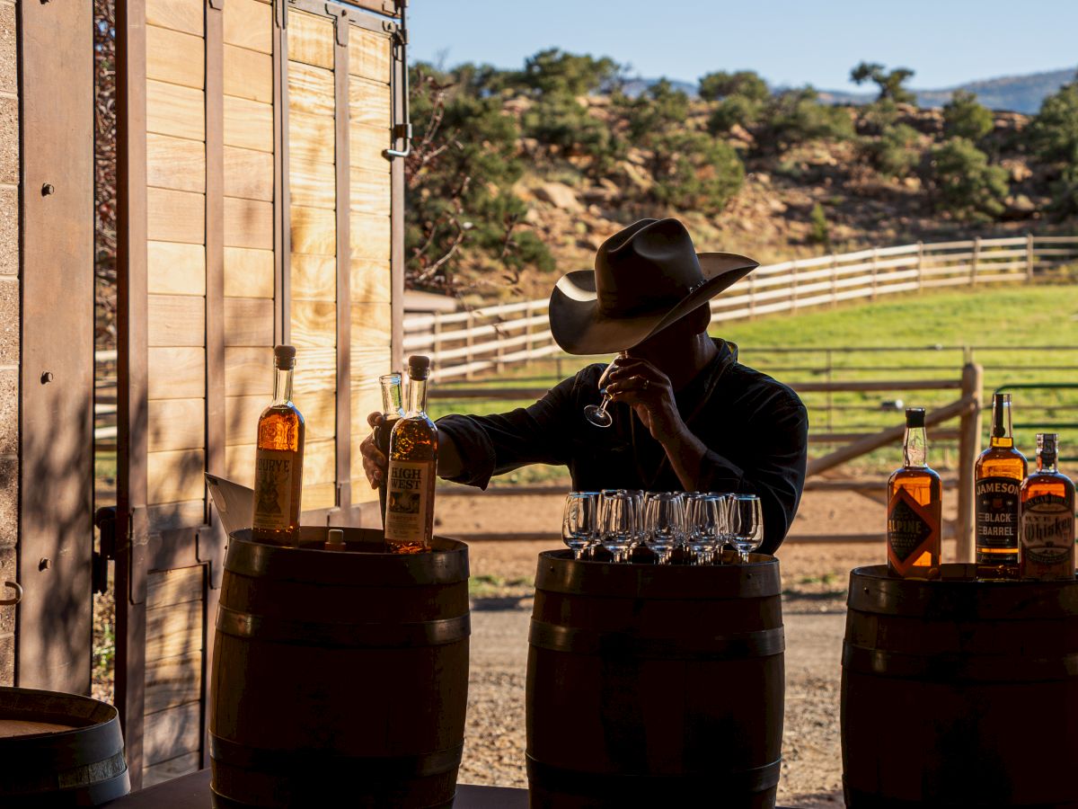 A person wearing a cowboy hat stands near barrels with bottles and glasses, set against a rural landscape with fencing in the background.